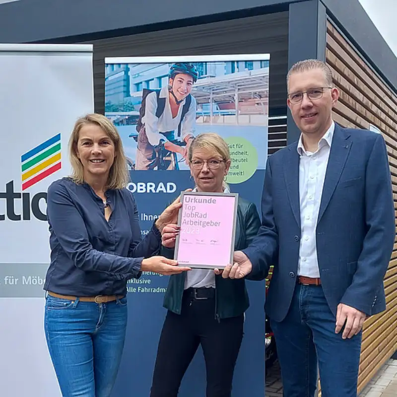 Monika Titze, von der Hettich Gruppe, steht zusammen mit einer Frau und einem Mann vor einem Poster mit dem Schriftzug Hettich und dem Hettich Logo. Sie alle tragen blaue Geschäftskleidung